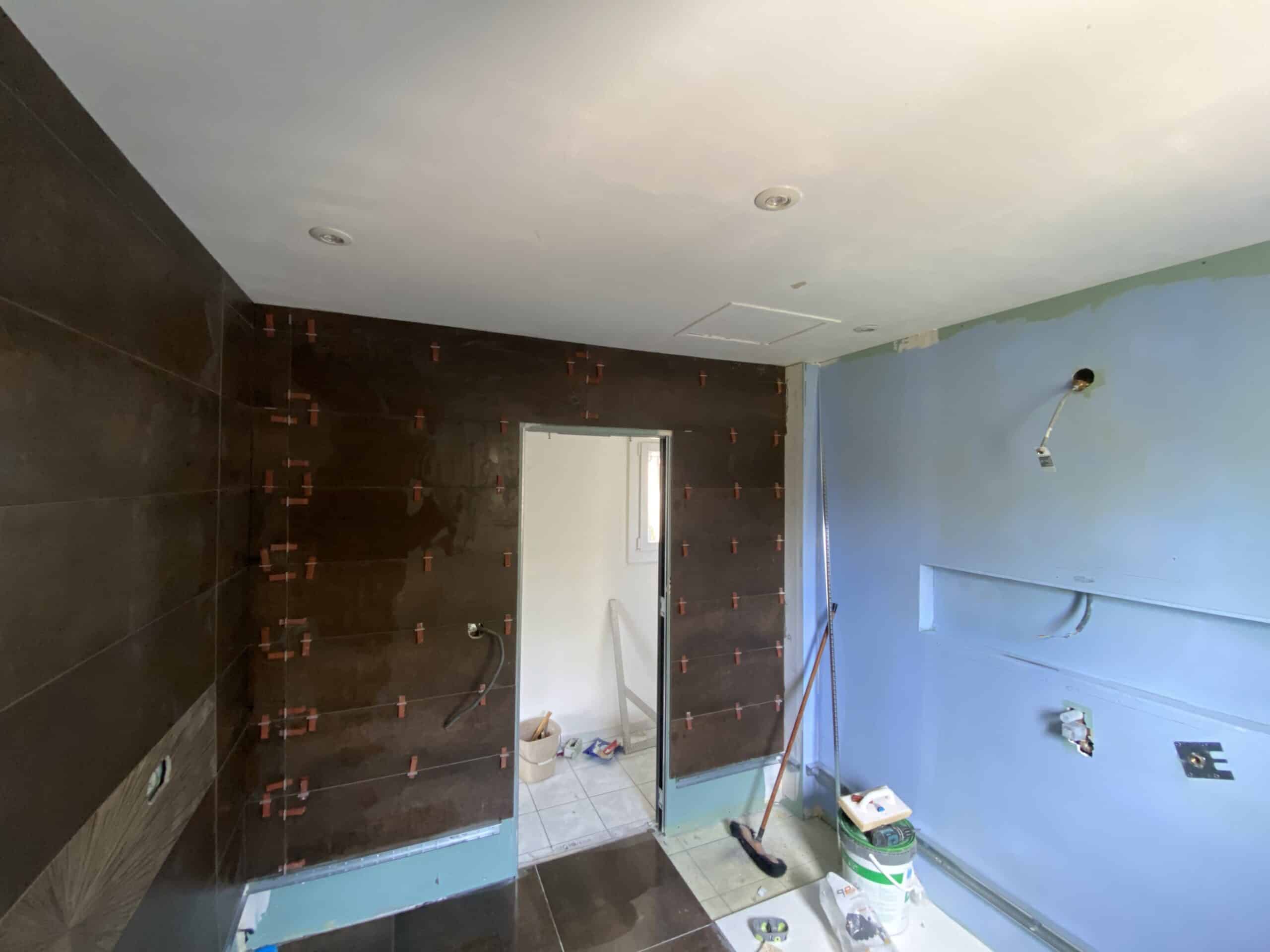 Rénovation de salle de bain : pose de grands carreaux muraux bruns foncés en cours (avec croisillons) face à une cloison bleue. Luminaires au plafond.