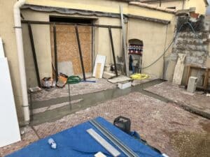 Chantier de rénovation extérieure : coulage de béton dans une saignée sur un sol en mosaïque, avec matériaux de construction et tuyaux.