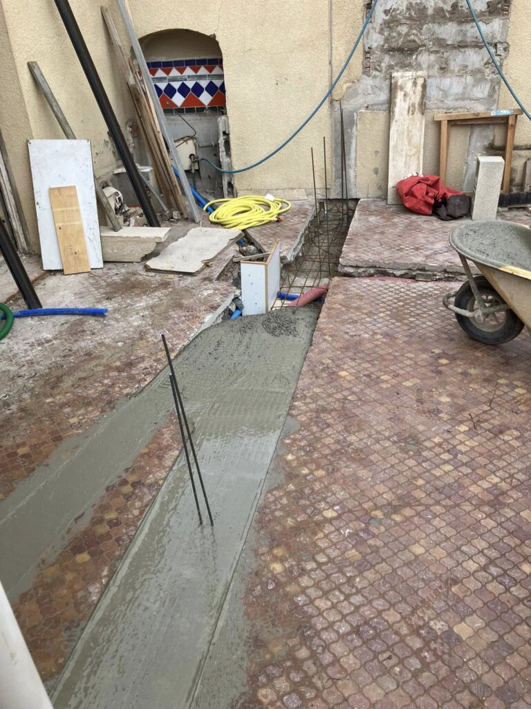 Coulage de béton sur terrasse pavée en rénovation Chantier extérieur : coulage de béton frais dans une tranchée, entouré de pavés rouges en forme d'écailles, avec matériaux et brouette.