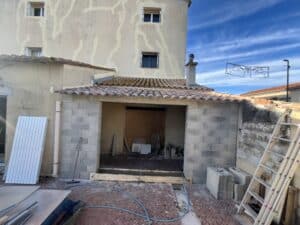 Construction d'une extension avec parpaings gris et tuiles romanes, devant un mur fissuré. Matériaux neufs et échelle sont visibles.