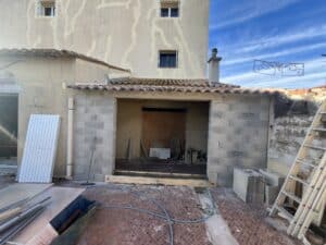 Construction d'une extension en parpaings sous un toit de tuiles, avec matériaux, tuyau bleu et façade fissurée CE OBRA visible.