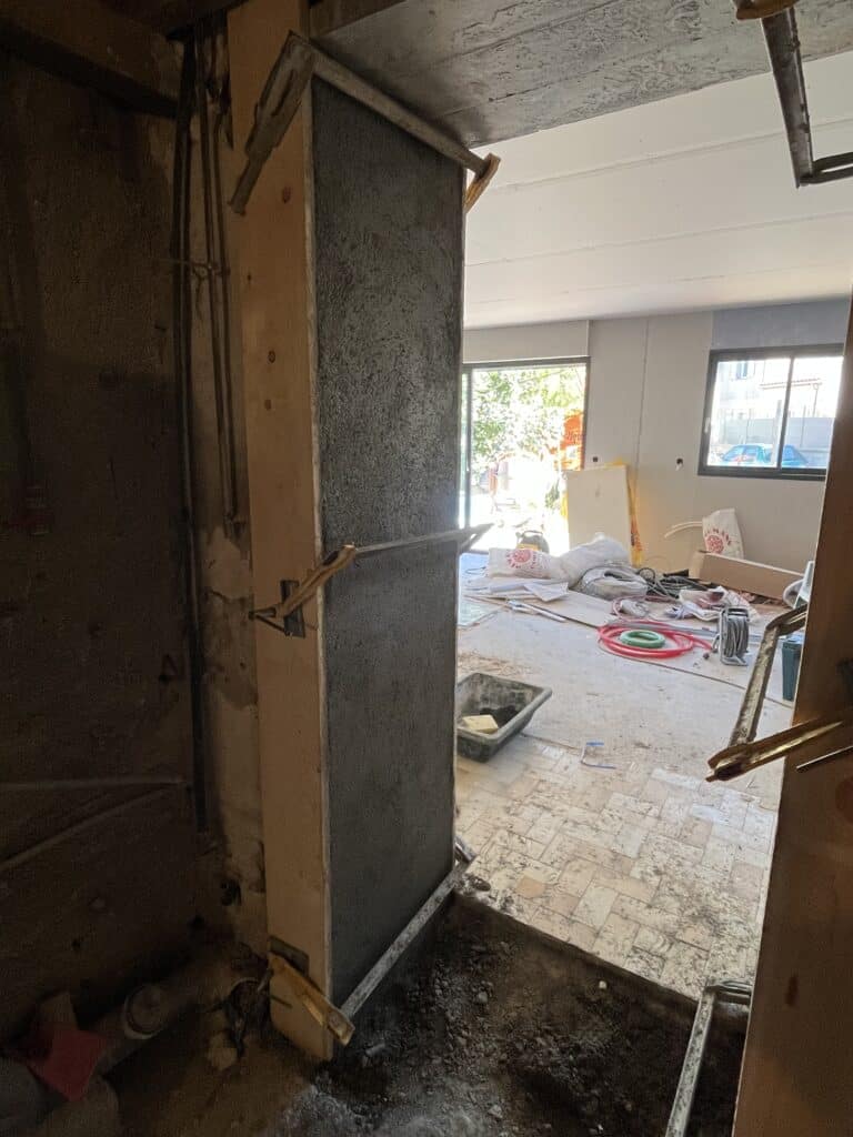 Chantier intérieur : mur de refend en cours de rejointoiement (chape rugueuse) soutenu par un étai métallique, vue vers une baie vitrée extérieure.