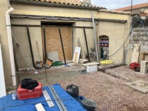 Chantier de rénovation extérieure avec matériaux (bâche bleue, tuyaux, métal) devant une façade beige et une ouverture murée en OSB. Sac 'BATI PLUS MAÇONNER' visible.