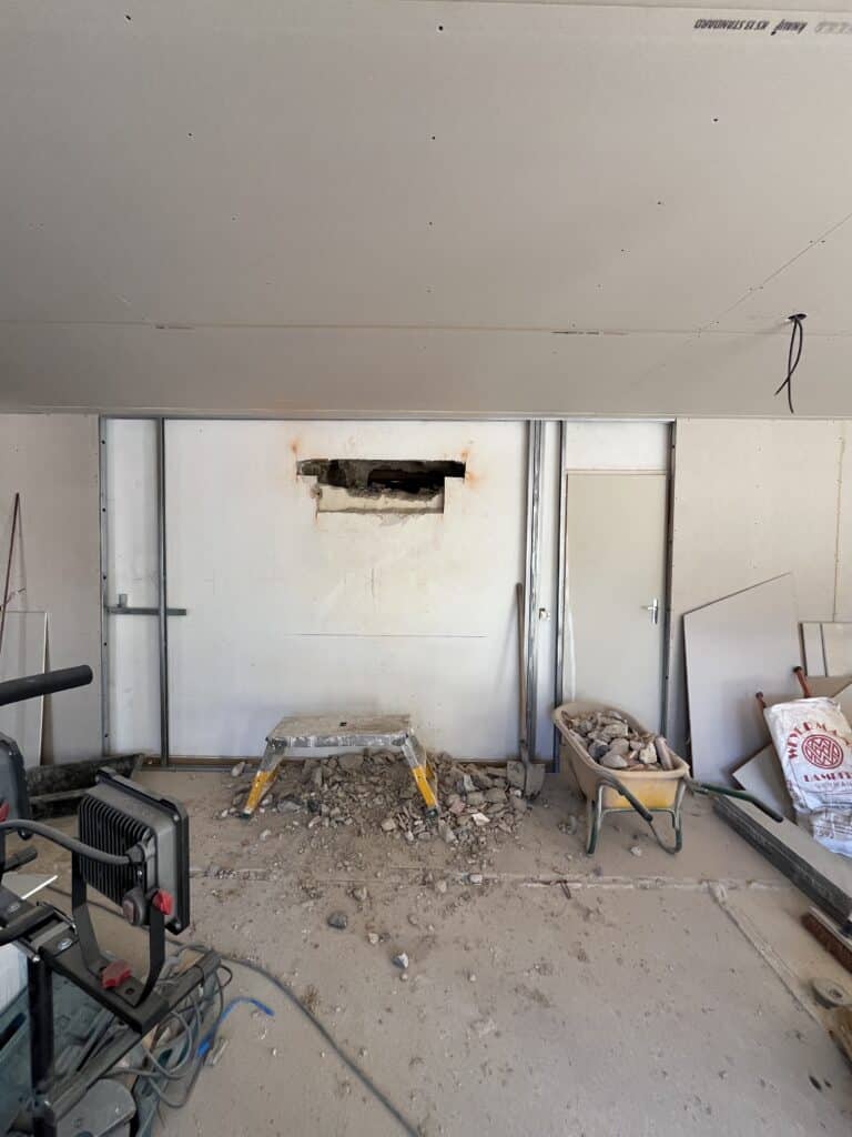 Chantier de rénovation avec mur en plaques de plâtre Knauf KS 13 STANDARD, trou de démolition, débris, tabouret, brouette et projecteur de travail.