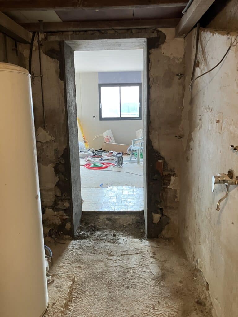Passage en rénovation avec murs bruts, chauffe-eau, et vue sur une pièce avec une fenêtre, des matériaux de construction (sacs 'EDMANN') et du carrelage neuf.