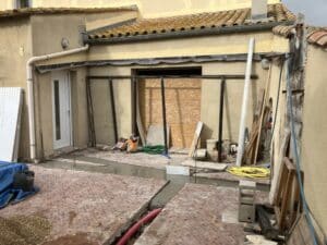 Chantier extérieur : sol en pavés rouges en cours de rénovation, tranchées de béton frais, matériaux de construction (bois, tuyaux, sacs de ciment).