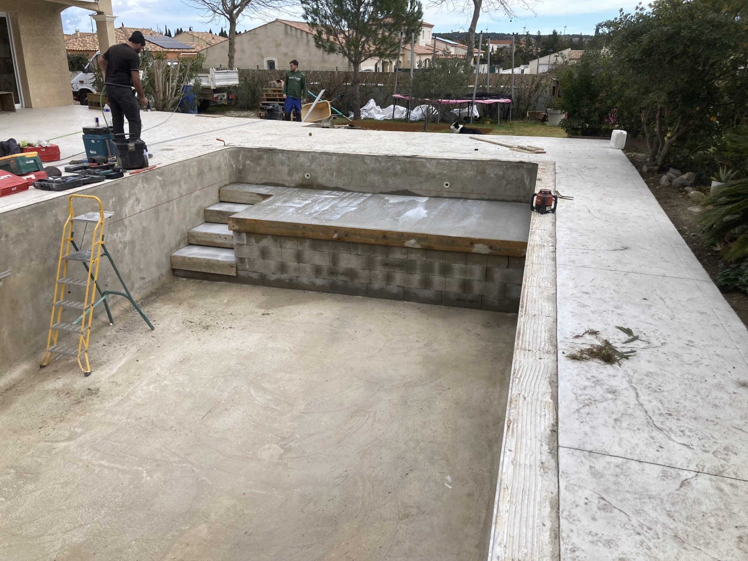 Piscine en béton en construction. Deux ouvriers aménagent les marches et la large plage immergée. Outils et échelle jaune visibles.