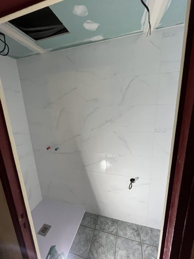 Douche en cours de rénovation : carrelage mural effet marbre blanc, receveur de douche blanc, évacuation visible, arrivées d'eau apparentes, et plafond en plaques de plâtre bleu clair.