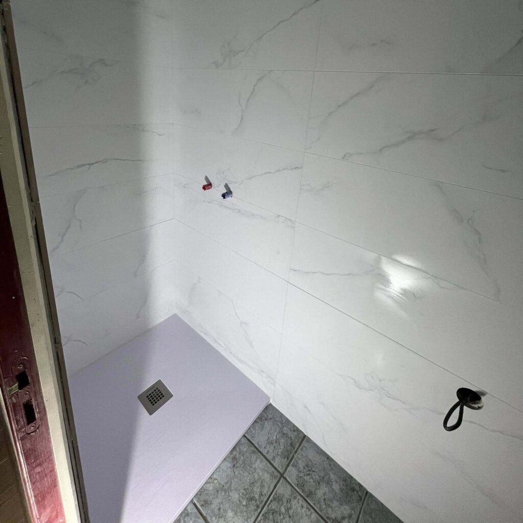 Douche en cours de rénovation, parois carrelées blanc marbré, receveur clair avec bonde carrée, sorties d'eau et support mural noir.