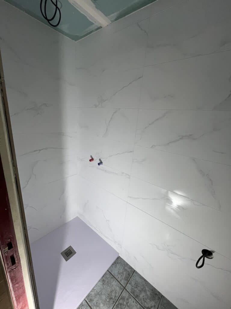 Douche en cours de rénovation : parois carrelées blanc marbré, receveur lilas avec bonde carrée, tuyaux apparents, sol gris foncé.