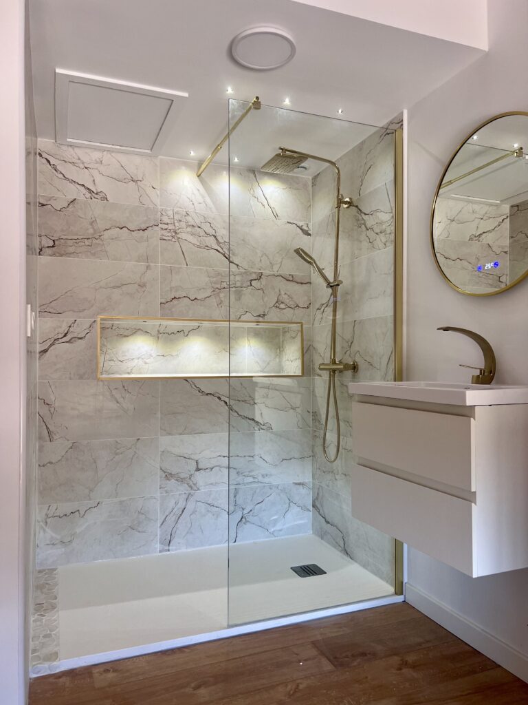Douche italienne luxueuse en marbre veiné rouge. Robinetterie laiton, niche illuminée et meuble vasque blanc avec miroir rond or.