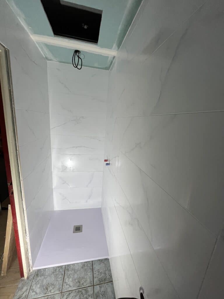 Douche en cours de rénovation avec carrelage mural imitation marbre blanc, receveur violet et évacuation, tuyauterie apparente au plafond.