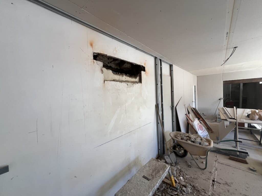 Chantier intérieur : mur en plaques de plâtre traversé par une ouverture irrégulière, structure métallique visible, matériel de construction (brouette, pelles) au sol.