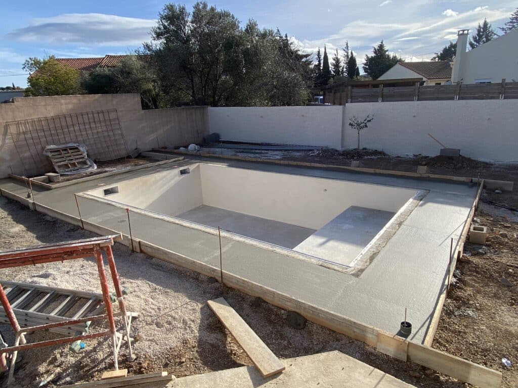 Bassin de piscine vide en construction, entouré de sa plage en béton fraîchement coulée. Chantier dans un jardin ensoleillé.