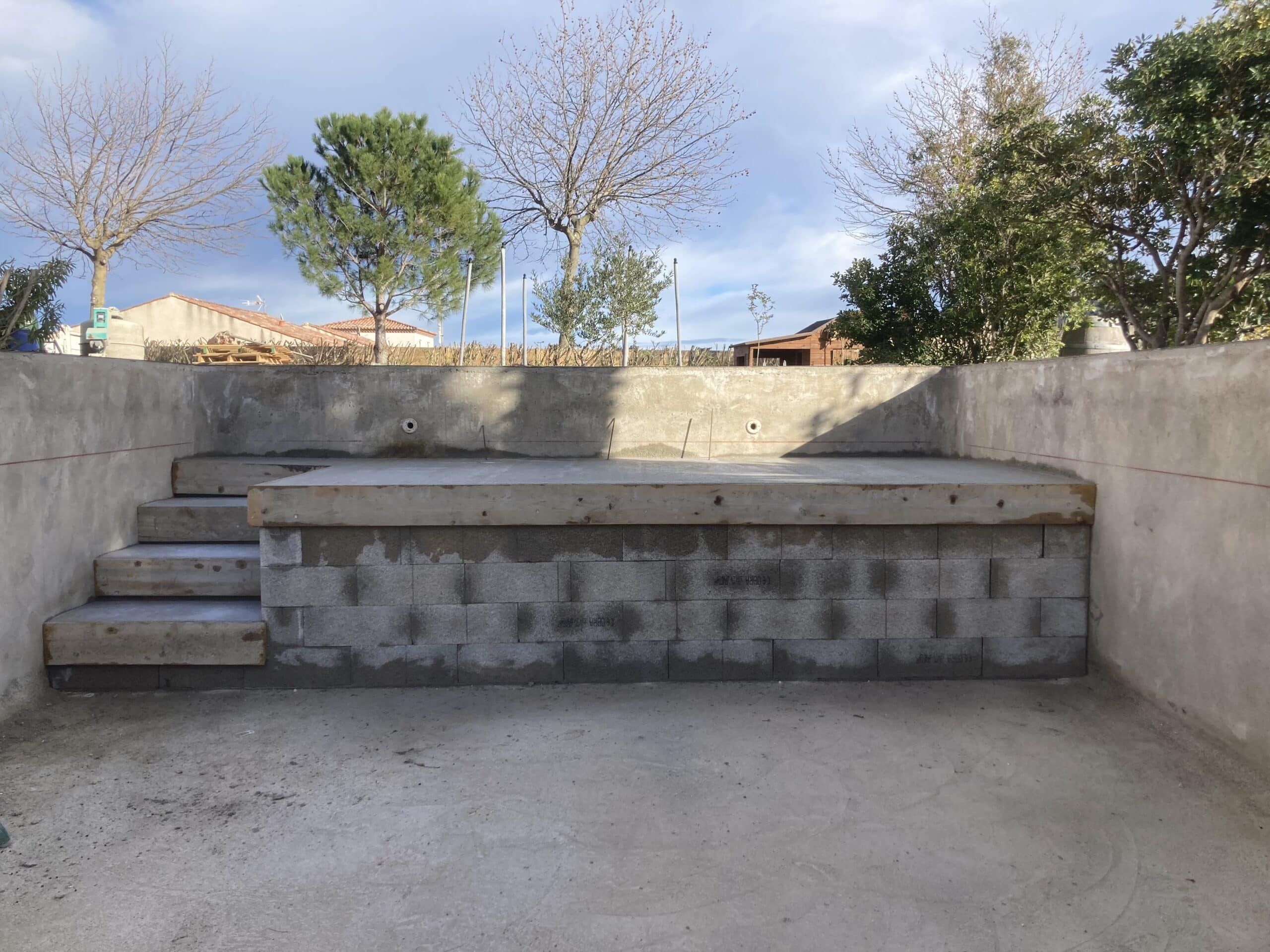 Construction d'une piscine en béton brut avec escalier d'angle et banquette centrale. Murs en blocs de parpaings et coffrage de bois.