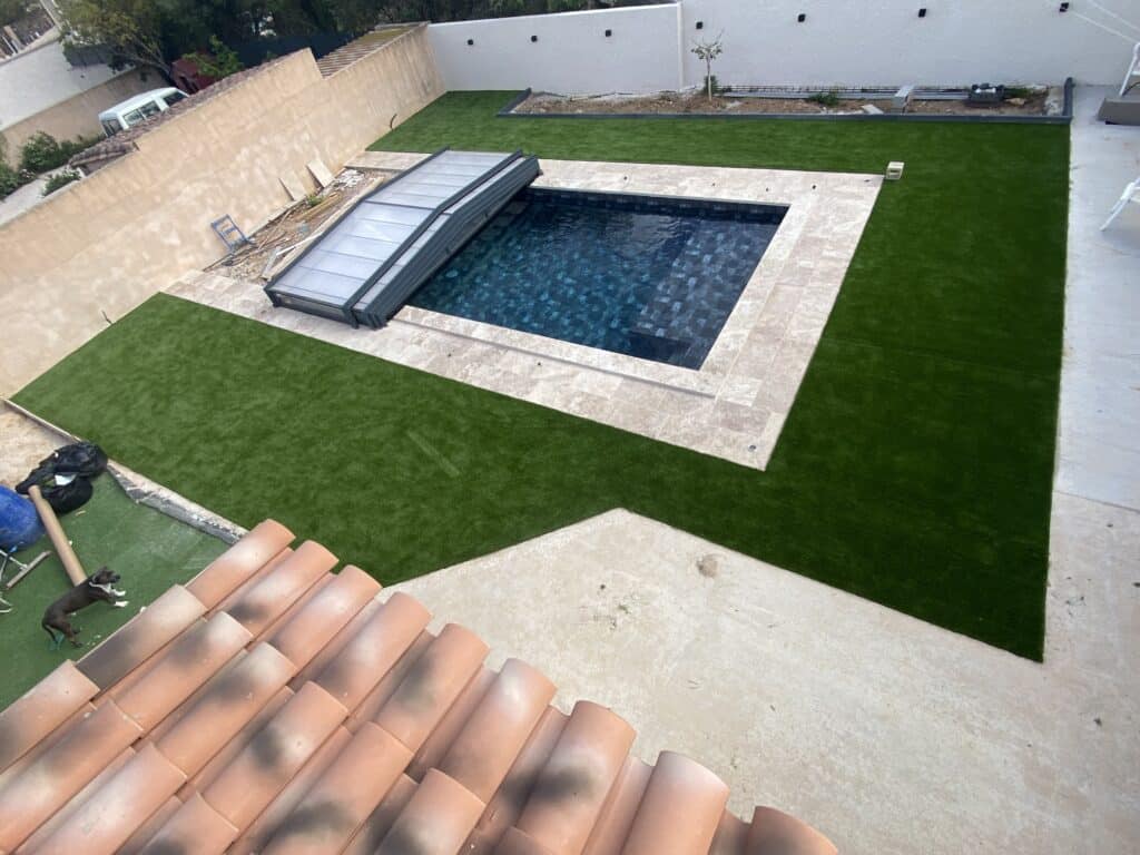 Aménagement moderne avec piscine à couverture automatique, plage en travertin clair et vaste jardin recouvert de gazon synthétique.