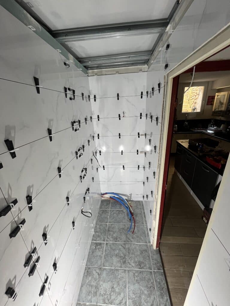 Chantier de salle de bain avec carrelage mural blanc marbré en cours de pose, utilisant des clips et des croisillons pour l'espacement. Tuyaux apparents au sol.