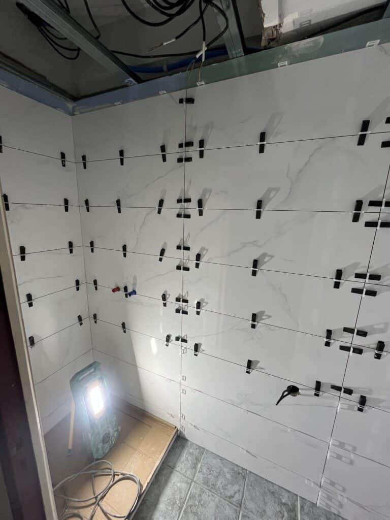 Pose de carrelage mural blanc marbré avec croisillons de nivellement noirs et fils tendus dans un coin, chantier éclairé par un spot.
