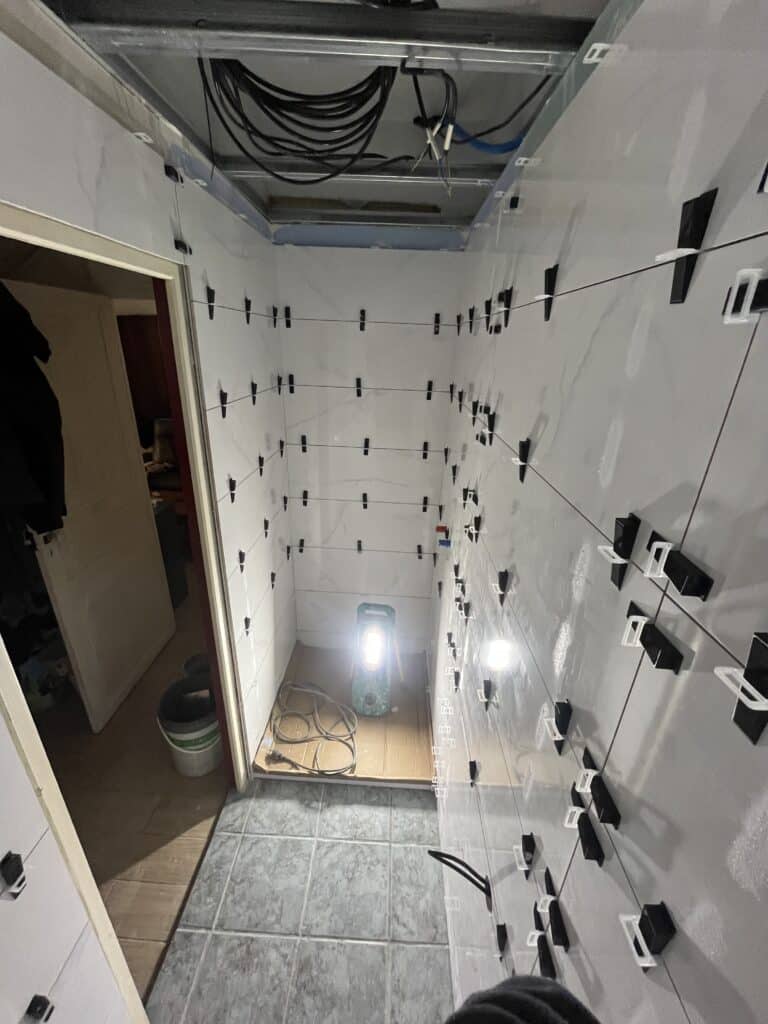 Pose de carrelage mural clair avec croisillons noirs et blancs et fils de nivellement tendus dans un espace confiné et sombre éclairé par un projecteur.