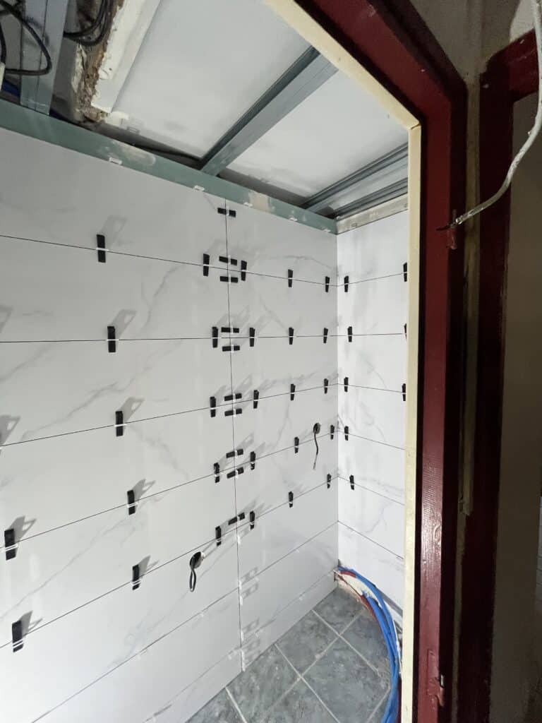 Pose de carrelage mural blanc effet marbre avec croisillons noirs et lignes de tirage, dans un angle de pièce en rénovation.