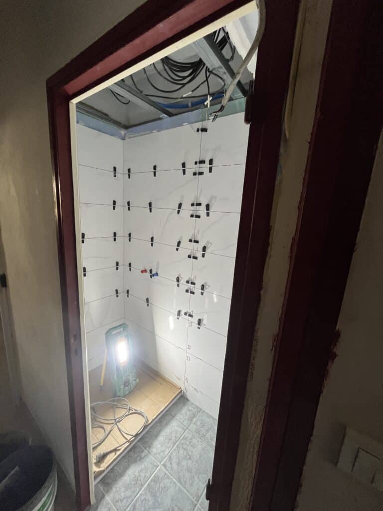 Pose de carrelage mural blanc dans une pièce sombre, vue à travers une porte avec chambranle rouge, avec niveaux et croisillons noirs en place.
