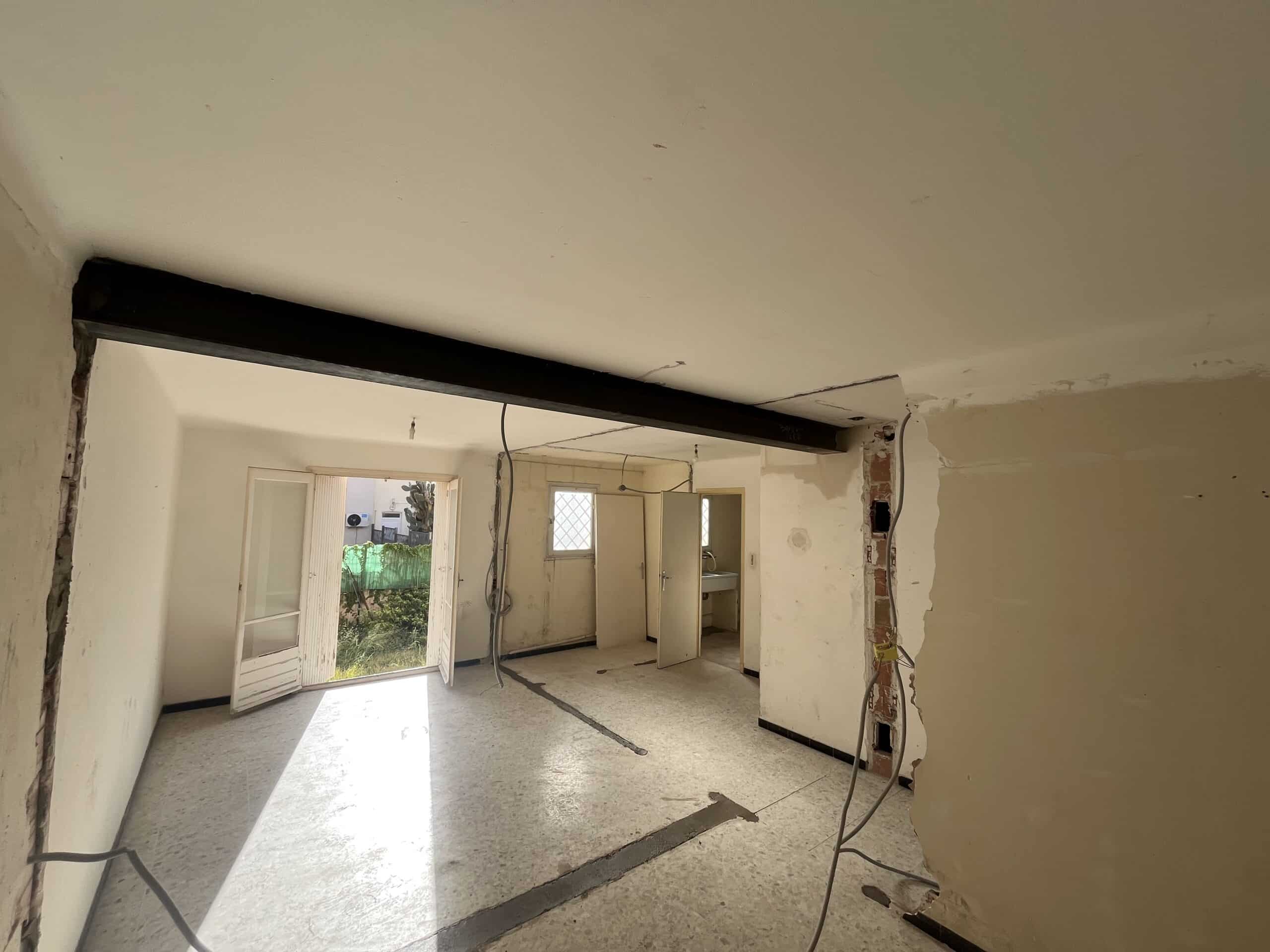 Intérieur en rénovation : une poutre en acier structurelle noire remplace le mur démoli. Câbles électriques et sol en terrazzo découpé. Portes ouvertes sur l'extérieur.