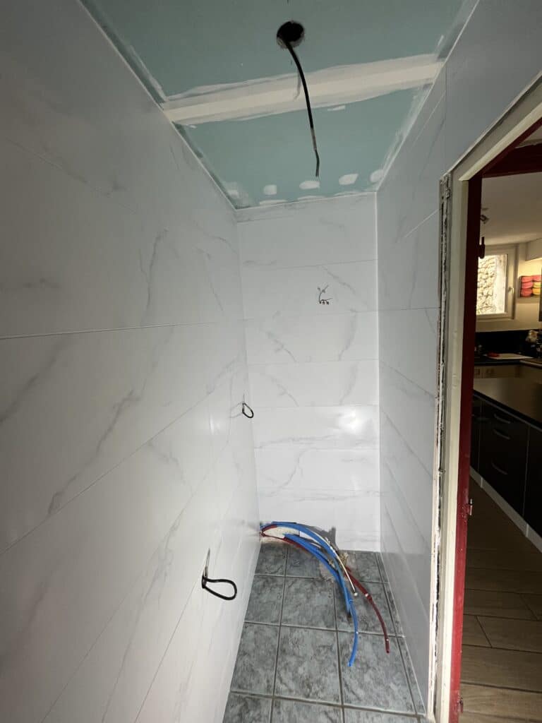 Douche en cours de rénovation : murs carrelés blanc marbré, sol gris, arrivée d'eau et câblage apparents, plafond en plaques de plâtre turquoise.