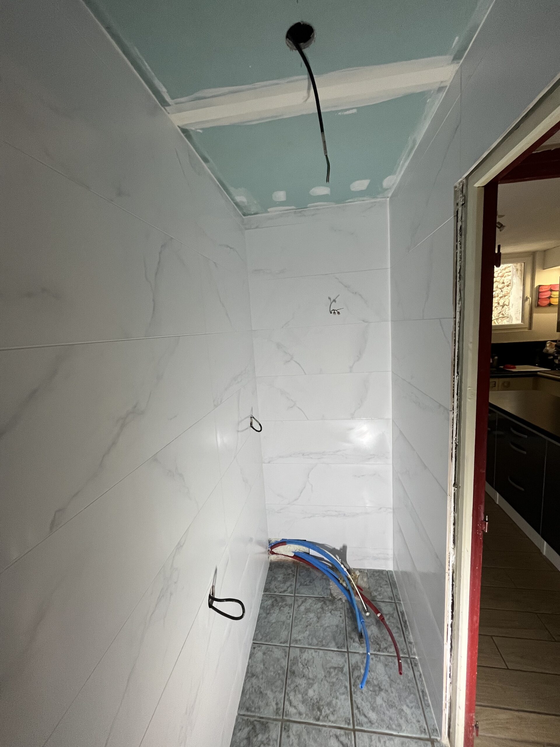 Douche en cours de rénovation : murs carrelés blanc marbré, sol gris, arrivée d'eau et câblage apparents, plafond en plaques de plâtre turquoise.
