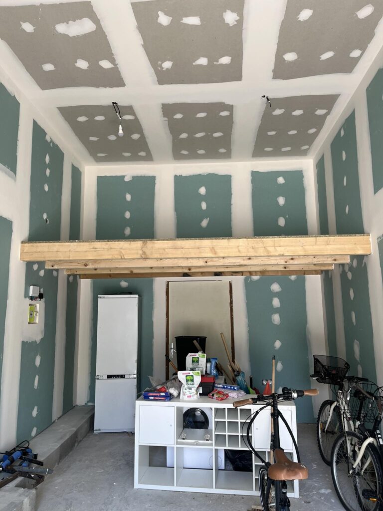 Rénovation Garage : Plaques Plâtre, Mezzanine et Vélos Garage en cours de rénovation avec plaques de plâtre jointoyées au plafond et sur les murs bleus/verts. Une mezzanine en bois brute est installée, entourée d'un réfrigérateur, d'un meuble cube blanc et de vélos.