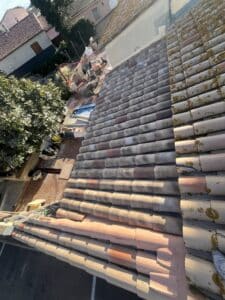 Vue plongeante sur des tuiles canal de toit en cours de rénovation, avec matériel de chantier, arbre et maisons voisines sous un ciel clair.