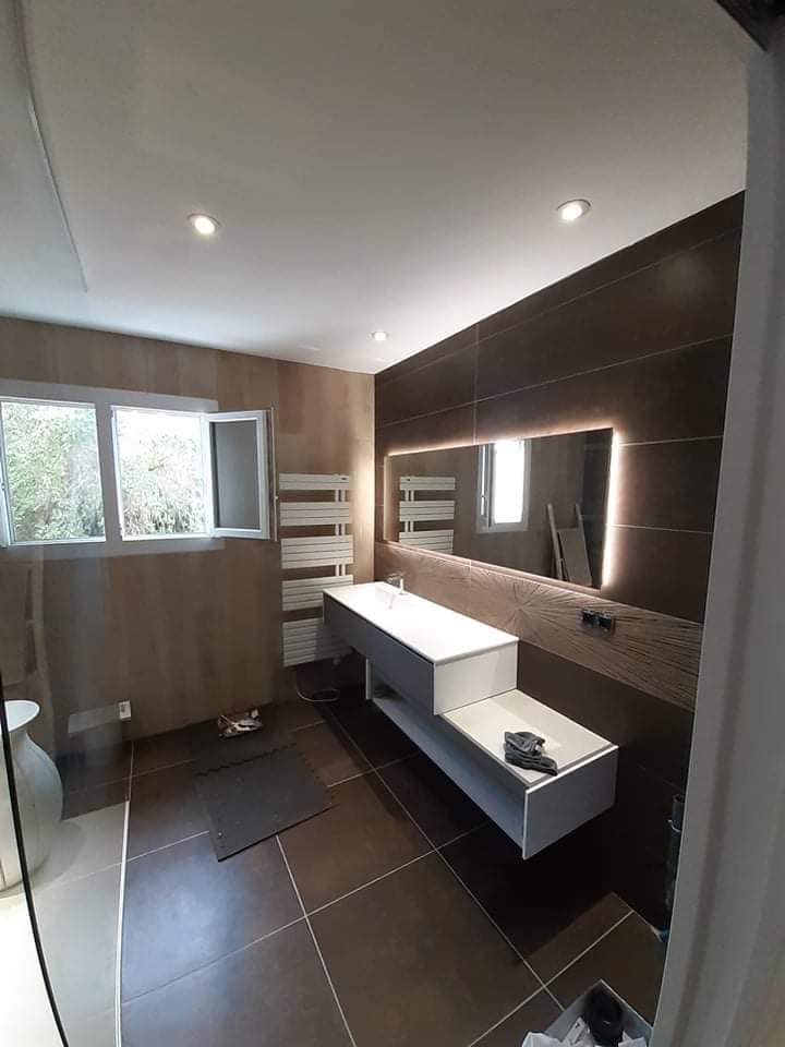 Salle de bain moderne : Lavabo suspendu et miroir rétroéclairé Salle de bain moderne avec carrelage marron foncé, meuble-lavabo suspendu blanc et grand miroir rétroéclairé. Fenêtres à gauche.