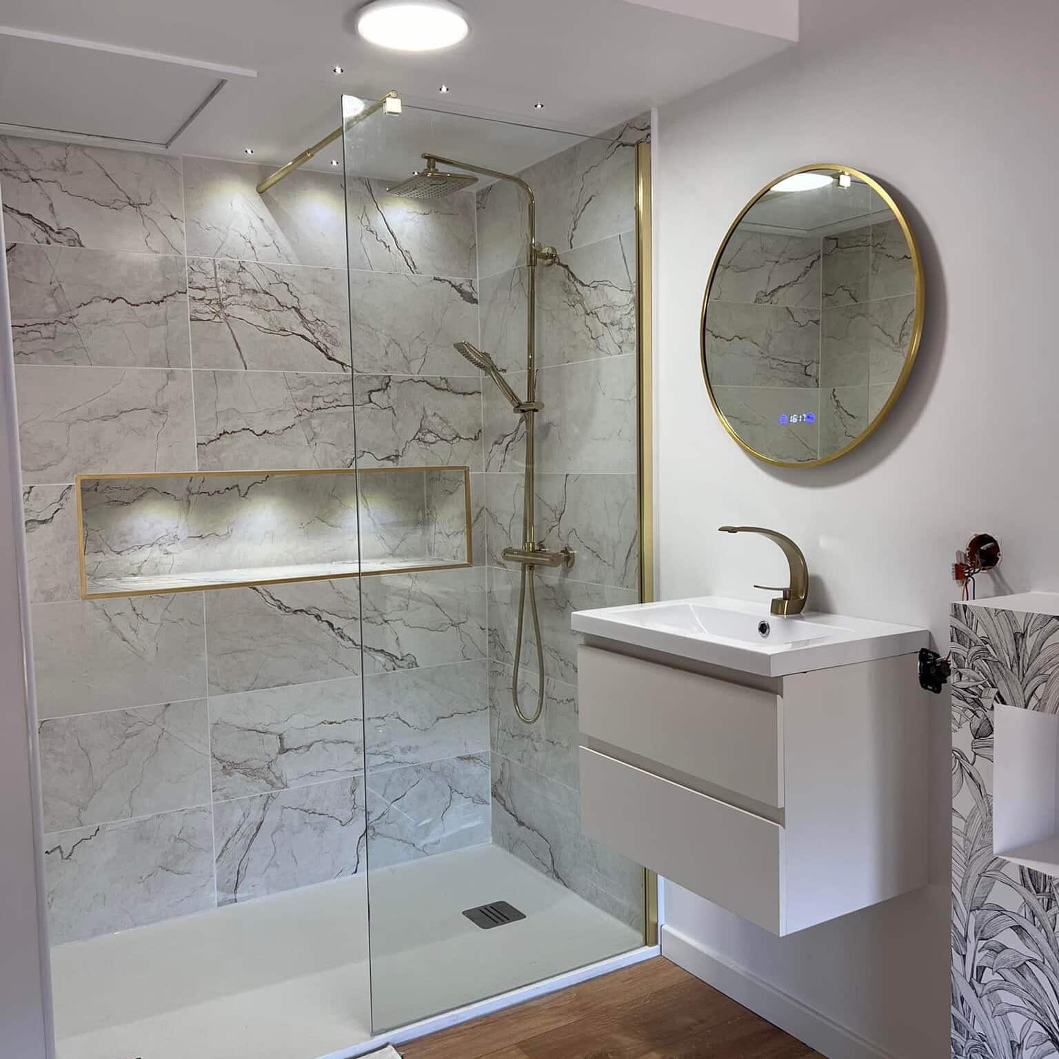 Salle de bain moderne avec douche carrelée effet marbre gris veiné, robinetterie et cadre miroir dorés, meuble-lavabo suspendu et miroir digital affichant 16:17.