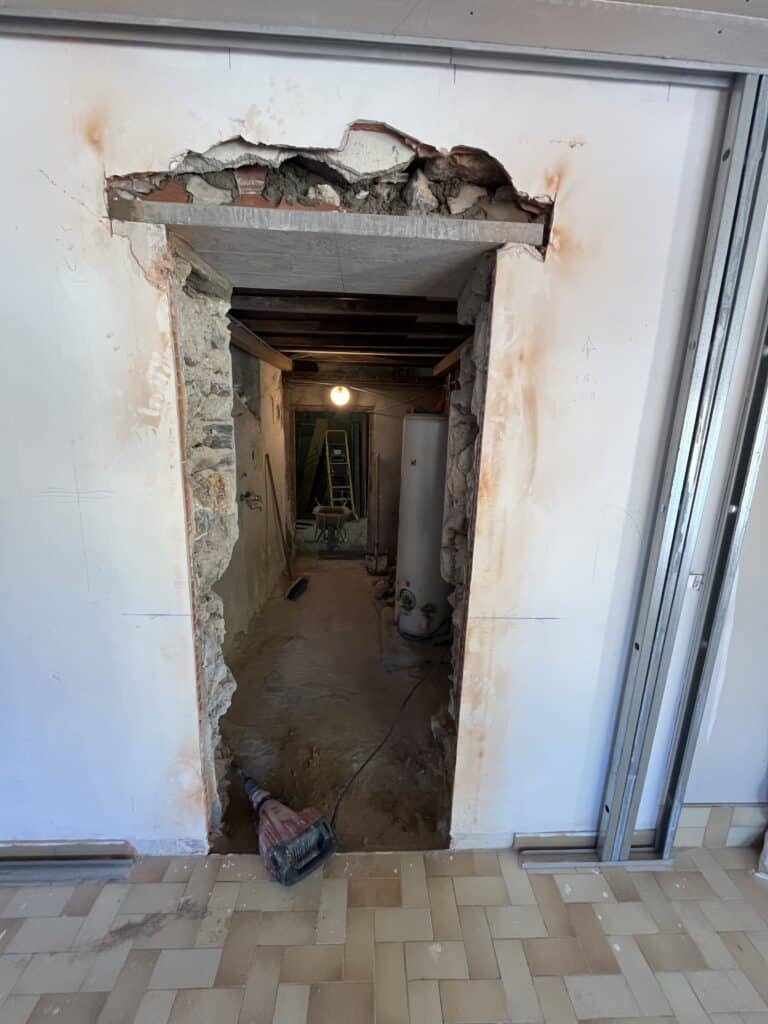 Ouverture de mur en cours de démolition montrant un linteau métallique, de la maçonnerie ancienne, et un marteau-piqueur rouge sur un sol carrelé.
