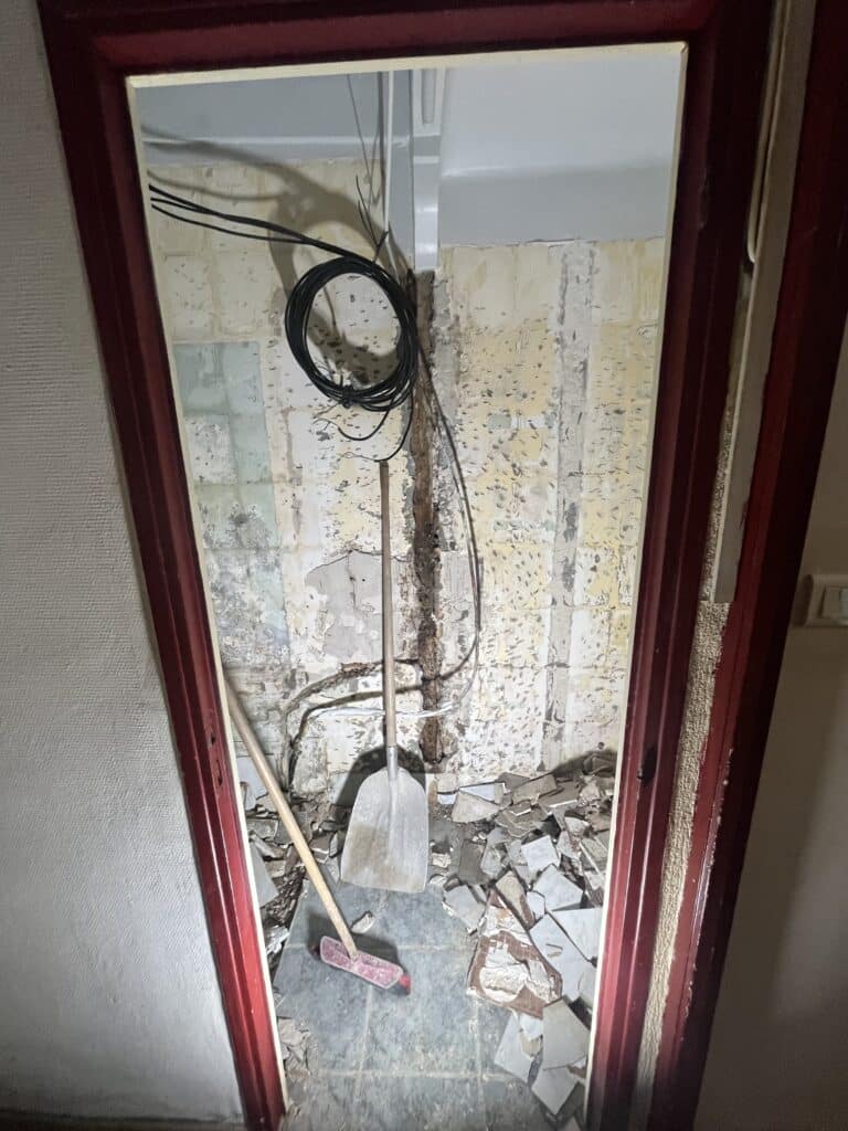 Porte entrouverte avec chambranle rouge, révélant des travaux de démolition : murs écaillés, câbles noirs enroulés, pelle et balai sur débris de carreaux cassés.