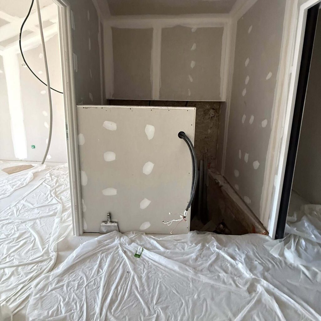 Travaux placo : jointoiement et préparation avant peinture Chantier de rénovation : mur en plaques de plâtre jointées, tuyaux apparents et sol protégé par une bâche plastique blanche.