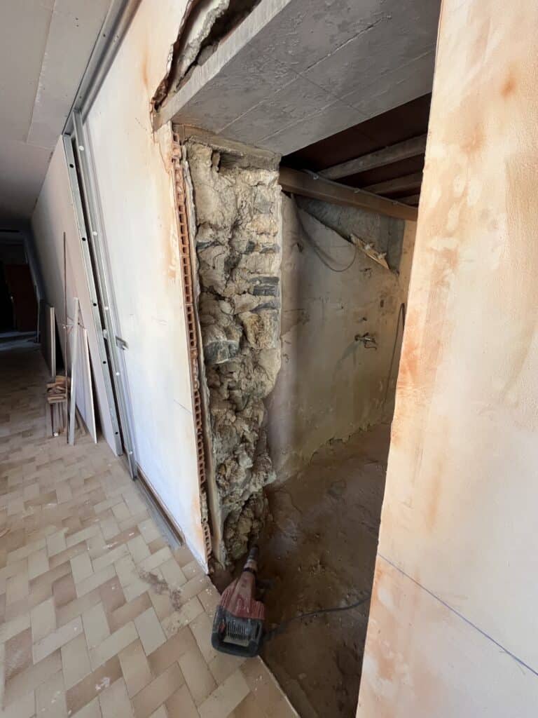 Travaux de rénovation : démolition d'une ouverture révélant un mur en pierres anciennes et un marteau-piqueur rouge au sol.